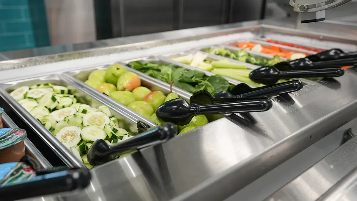 Salad bar with metal trays of sliced cucumbers, apples, leafy greens, celery, and other vegetables with serving tongs.